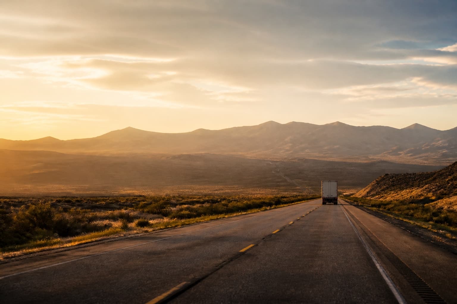 A box truck far ahead on a two-lane highway crossing high desert toward mountains at sunset