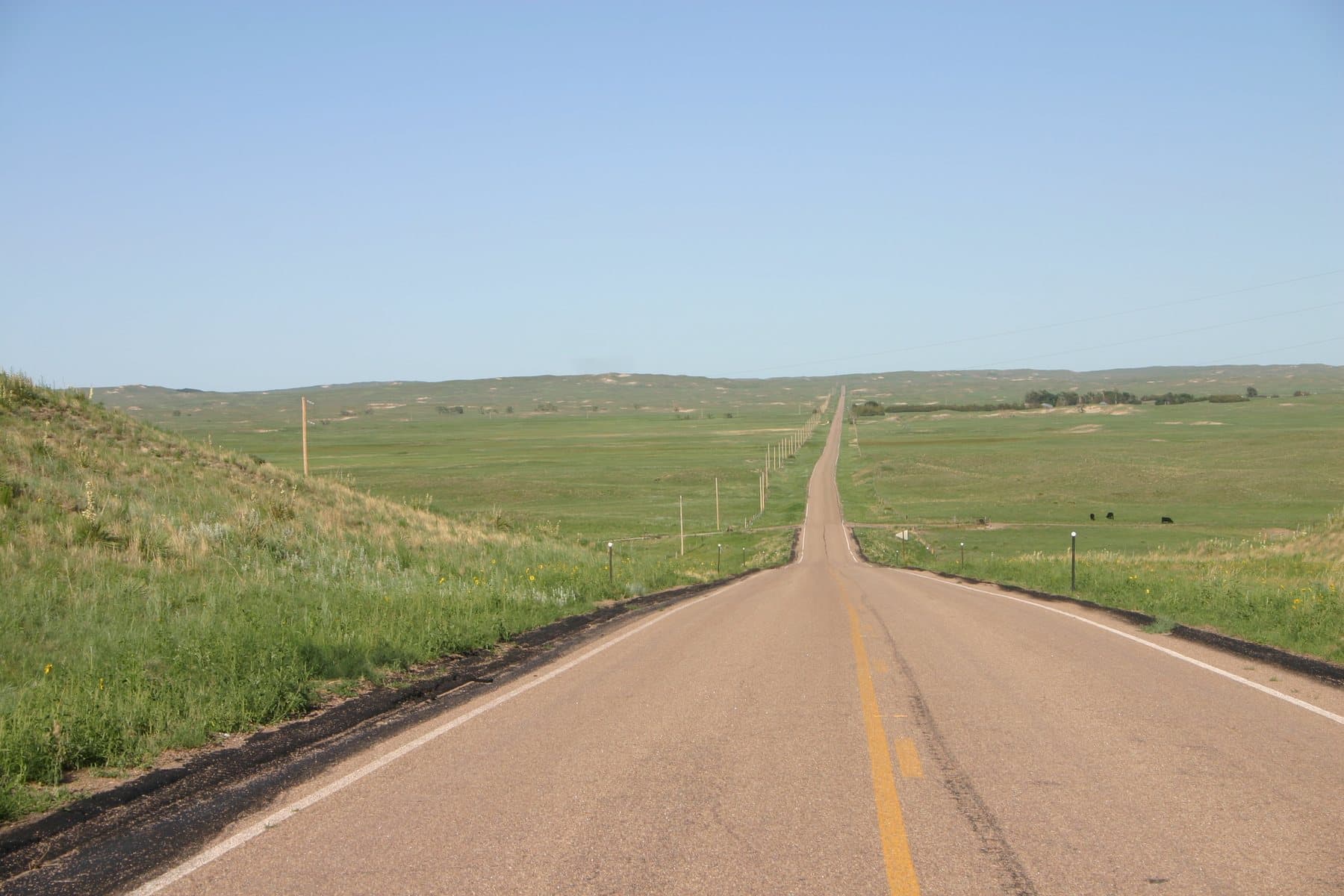 A straight two-lane highway running to the horizon across green Sandhills grassland under a clear blue sky